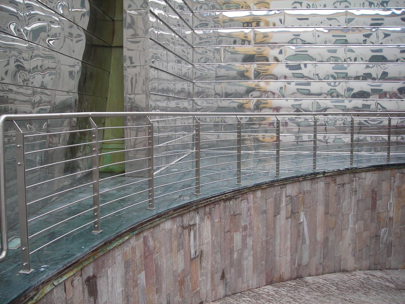Parapetto in acciaio inox lungo una rampa, con parete in lamiera lavorata a effetto specchio
